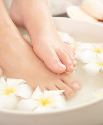 closeup view of woman soaking her feet in dish with water and flowers on wooden floor. Spa treatment and product for female feet and hand spa. white flowers in ceramic bowl.